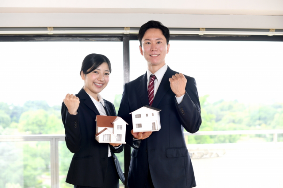 不動産投資をイメージしたスーツ姿の男女。住宅模型を手に持ち、笑顔でガッツポーズをしている様子。
